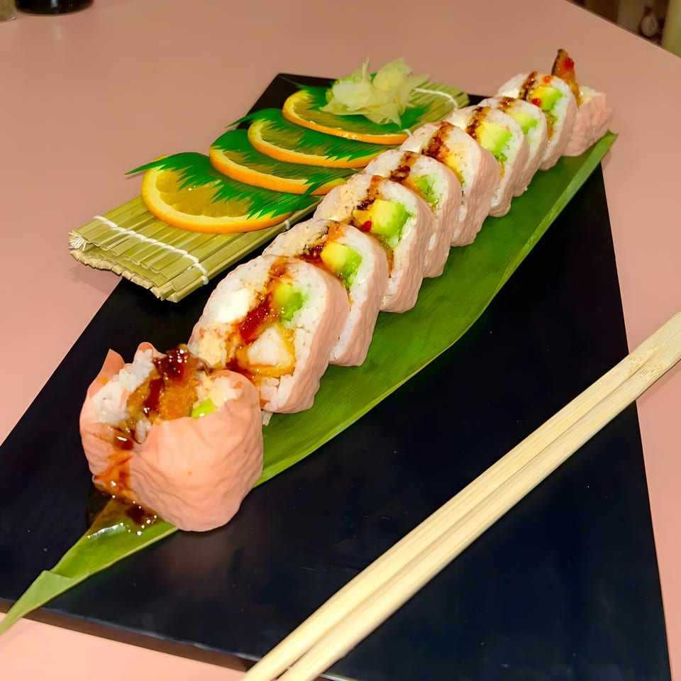 Pink sushi rolls with avocado and sauce, garnished with orange slices and ginger, on a black plate.