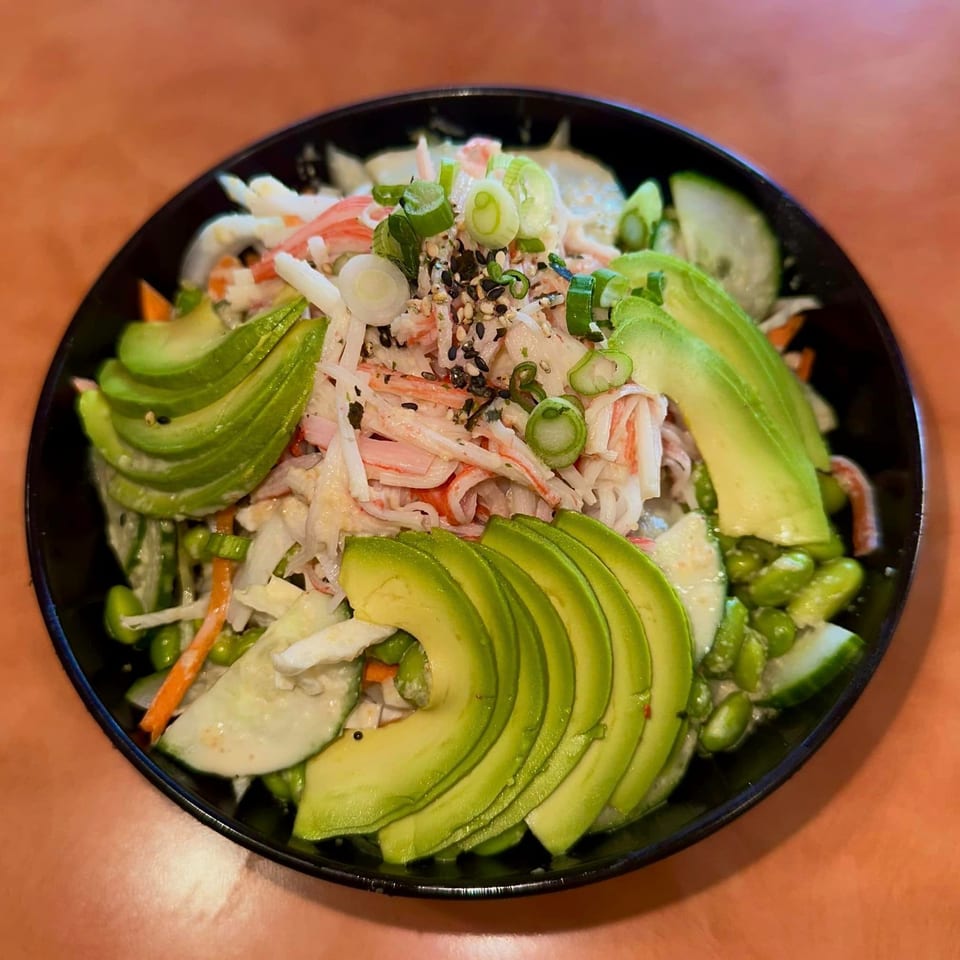 A black bowl filled with an Asian-fusion salad, featuring crab, avocado, cucumber, and edamame.